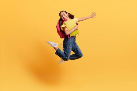 Photo for Happy cute preteen girl in casual with backpack and books notepads in her hands jumping up, smiling and gesturing isolated on yellow background, thrilled kid going to school - Royalty Free Image