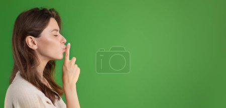 Photo for Serious pretty young european lady in casual puts finger to lips, making secret gesture isolated on green studio background, profile. Ad and offer, mystery, surprise, panorama - Royalty Free Image