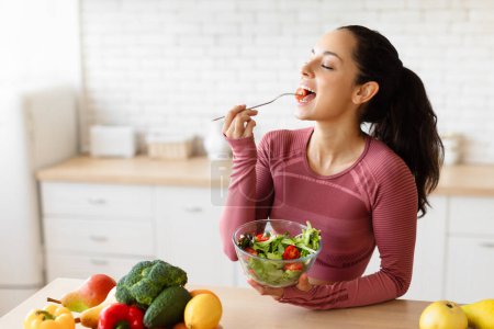 Photo for Tasty Weight Loss. Fit lady eating fresh salad holding bowl, enjoying vegetable meal at modern kitchen table indoors, wearing fitwear. Healthy Slimming Diet And Sport Nutrition - Royalty Free Image