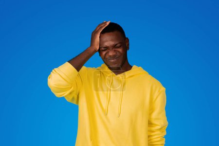 Photo for Sad young black guy presses hand to head, suffers from mistake and headache, isolated on blue background, studio. Human emotions, migraine, disease, health problems and stress - Royalty Free Image