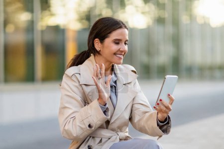 Photo for Happy attractive mature businesswoman in formal outfit beige trench coat have video call, waving and smiling at smartphone screen, sit on bench on street, have online meeting with family, copy space - Royalty Free Image