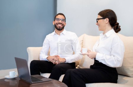 Photo for Business Communication. Smiling Businesswoman Conducting a Job Interview with a New Employee Man, Sitting On Sofa Indoor. Sipping Coffee And Laughing Enjoying Productive Day - Royalty Free Image