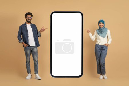 Photo for Cheerful Arabic Couple Pointing At Big Blank Smartphone With White Screen, Happy Muslim Man And Woman In Hijab Advertising New Mobile App Or Website, Posing On Beige Background, Mockup, Collage - Royalty Free Image