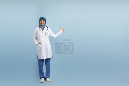 Téléchargez les photos : Publicité médicale. Heureux médecin musulmane pointant de côté à l'espace de copie, médecin arabe dame portant uniforme debout sur fond bleu studio, pleine longueur avec place vide pour la conception - en image libre de droit