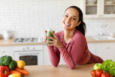 Photo for Sports Nutrition. Smiling young arabic lady in sportswear at table holds glass with fresh smoothie in minimalist kitchen interior, looking at camera. Healthy weight loss diet, detox and body care - Royalty Free Image