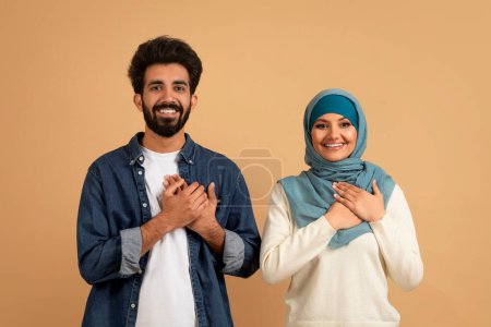 Téléchargez les photos : Couple musulman reconnaissant gardant les deux mains sur la poitrine, exprimant gratitude et appréciation, gentil homme arabe et femme en hijab ayant de bons sentiments, debout sur fond de studio beige, espace libre - en image libre de droit