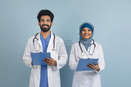 Photo for Portrait of happy arab male and female doctors in uniform holding clipboards and smiling at camera, muslim therapist man and woman with stethoscopes posing over blue studio background, free space - Royalty Free Image