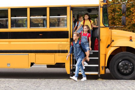 Foto de Concepto de transporte. Grupo de niños felices saliendo del autobús escolar amarillo, diversas colegialas sonrientes y colegiales saliendo del vehículo, listos para las lecciones, disfrutando del estudio juntos - Imagen libre de derechos