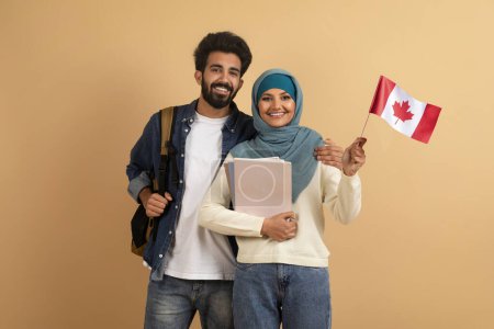Téléchargez les photos : Couple arabe souriant avec drapeau canadien et cahiers d'exercices entre les mains posant sur fond de studio beige, heureux musulmans hommes et femmes étudiants bénéficiant de cours de langues étrangères et étudier à l'étranger, espace libre - en image libre de droit