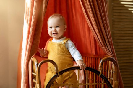 Téléchargez les photos : Gros plan de bébé adorable debout dans son berceau rétro en bois à la maison, drôle mignon petit tout-petit garçon portant une combinaison souriante et regardant loin, se détendre au lit à l'intérieur, Copier l'espace - en image libre de droit