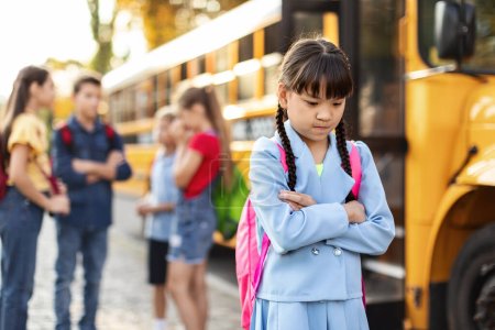 Photo for Sad little asian schoolgirl standing alone near school bus while her classmates chatting on background, upset preteen female child suffering social exclusion problem, feeling lonely and depressed - Royalty Free Image