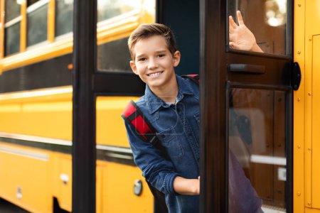 Foto de Feliz niño preadolescente de pie en la puerta del autobús escolar y mirando a la cámara, alegre alumno masculino con mochila asomándose desde el autobús escolar amarillo, listo para el viaje a casa después de las lecciones, espacio para copiar - Imagen libre de derechos