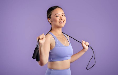 Foto de Entrenamiento cardiovascular. Sonriente mujer asiática en ropa deportiva sosteniendo saltar la cuerda alrededor del cuello, escuchando música en auriculares mientras posando en el estudio sobre fondo púrpura - Imagen libre de derechos