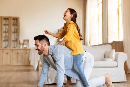Photo for Funny Family Playtime. Young arabic father riding his daughter on back, fooling and playing at home, kid laughing while playing cowgirl on horse with her dad at modern living room interior - Royalty Free Image