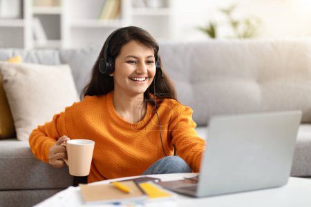 Photo for Happy beautiful millennial hindu woman remote employee have online work meeting with colleagues, sitting on floor at home, using headset, drinking coffee and smiling at laptop camera, copy space - Royalty Free Image