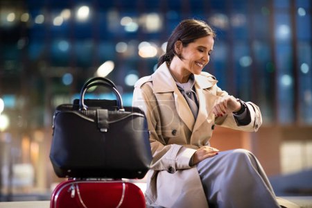 Téléchargez les photos : Femme d'affaires assise au salon de l'aéroport, attendant le vol. Bonne femme d'âge moyen assis à l'aéroport salle d'attente avec des bagages, regardant la montre sur son poignet, l'heure d'enregistrement. Voyages d'affaires - en image libre de droit