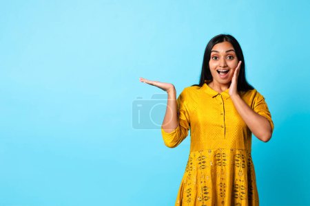 Photo for Look At This Offer. Excited Middle Eastern Woman In Yellow Traditional Dress Advertising Text, Holding Invisible Product, Showing Empty Space For Text Over Blue Studio Background - Royalty Free Image