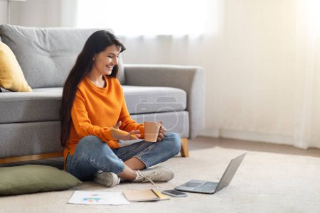 Photo for Positive friendly millennial indian woman remote employee have video conference with colleagues. Young lady sit on floor in living room at home, hold coffee mug, looking at laptop screen, copy space - Royalty Free Image