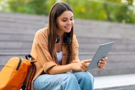 Photo for Cool pretty long-haired brunette young woman in stylish casual outfit student sitting at park with backpack and wireless headphones, using digital tablet, checking her schedule, empty space - Royalty Free Image