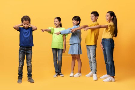 Photo for Offended upset preteen boy suffering from bullying from children. Laughing diverse kids pointing at guy covering his ears, isolated on yellow background. Bullying problem among school children - Royalty Free Image