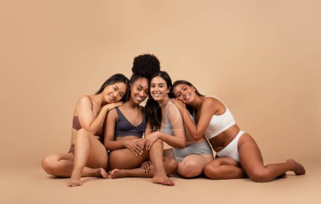 Foto de Grupo de jóvenes mujeres internacionales en diferentes calzoncillos con belleza natural, sentadas en el suelo sobre fondo beige estudio. Poder femenino, salud y feminismo - Imagen libre de derechos