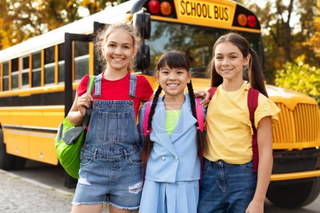 Photo for Portrait Of Three Happy Girls Posing Outdoors Near School Bus Together, Cheerful Preteen Multiethnic Female Friends With Backpacks Standing Outside, Classmates Embracing And Smiling At Camera - Royalty Free Image