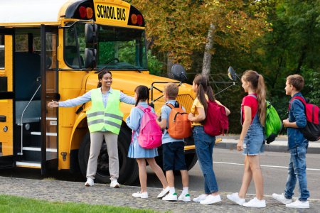 Foto de Maestra negra ayudando a los niños mientras entran en el autobús escolar, la asistente afroamericana en el chaleco reflectante mostrando el camino a la escuela, grupo de niños guía con mochilas - Imagen libre de derechos
