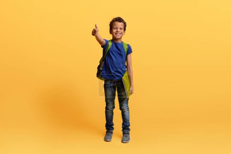 Téléchargez les photos : Joyeux écolier noir pré-adolescent mignon avec sac à dos et carnet d'exercices montrant pouce levé et souriant isolé sur fond de studio jaune. Enfant afro-américain joyeux allant à l'école, copier l'espace - en image libre de droit