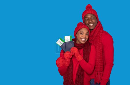 Foto de Viajes de vacaciones de invierno. Feliz joven pareja negra sosteniendo pasaportes con boletos de tarjeta de embarque, sonriendo a la cámara posando en cálidas bufandas rojas y sombreros en el fondo del estudio azul, espacio de copia - Imagen libre de derechos