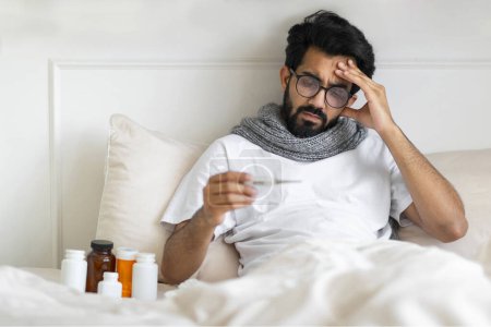 Photo for Sick Indian Man Wearing Scarf Sitting In Bed And Holding Thermometer, Young Ill Eastern Guy Measuring Body Temperature, Having Flu Symptoms And Touching Forehead In Frustration, Free Space - Royalty Free Image