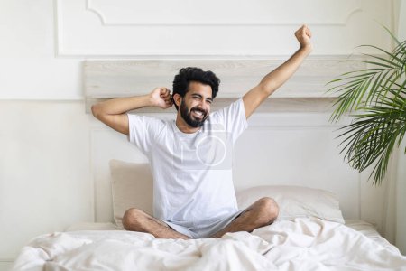 Photo for Good Morning. Happy Indian Guy Sitting On Bed And Stretching Arms After Good Sleep, Smiling Handsome Young Eastern Man Relaxing In Cozy Bedroom, Cheerful Male Enjoying Home Leisure, Copy Space - Royalty Free Image