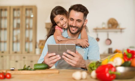Téléchargez les photos : Recettes en ligne. Père heureux et sa fille en utilisant une tablette numérique, fille embrassant papa du dos tout en cuisinant la salade ensemble dans la cuisine, panorama, espace libre - en image libre de droit
