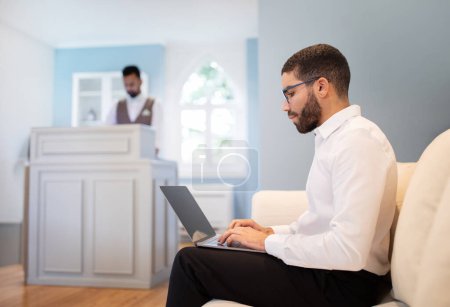 Foto de Viajes de negocios. Hombre viajero de negocios de Oriente Medio que trabaja en línea en la computadora, sentado en el sofá cómodamente en el vestíbulo del hotel interior, participando en el trabajo de Internet, escribiendo en el ordenador portátil, enfoque selectivo - Imagen libre de derechos