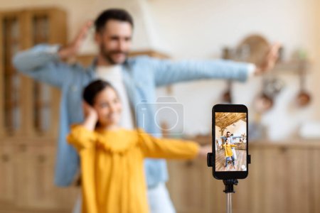 Téléchargez les photos : Médias sociaux Fun. Cheerful Middle Eastern Father And Daughter Influencers Tournage vidéo de danse sur téléphone portable à la maison, la création de contenu pour la famille en ligne Vlog. Focus sélectif sur l'écran du smartphone - en image libre de droit