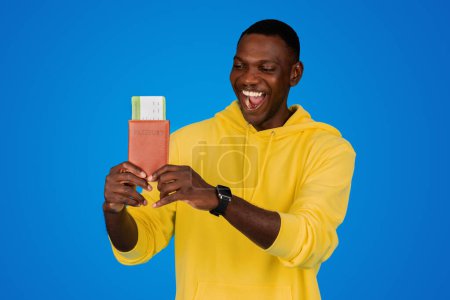 Photo for Smiling young black guy with open mouth looks at ticket and passport, shouts and rejoices to journey, isolated on blue background, studio. Emotions, victory, trip, tourism and vacation - Royalty Free Image