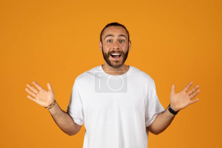 Photo for Smiling surprised caucasian millennial man with beard in white t-shirt spreads arms to sides, isolated on orange studio background. Victory, good news, human emotions, ad and offer, lifestyle - Royalty Free Image