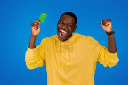 Téléchargez les photos : Riant heureux excité jeune homme noir montre carte de crédit, se réjouit de la victoire, célèbre le succès, isolé sur fond bleu, studio. Émotions humaines, publicité et offre, profit et shopping - en image libre de droit