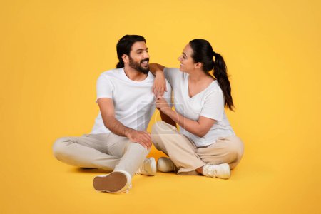 Photo for Happy millennial arab guy and woman in casual talk, sit on floor, enjoy communication, spare time together, isolated on yellow studio background. Family and relationships, lifestyle - Royalty Free Image