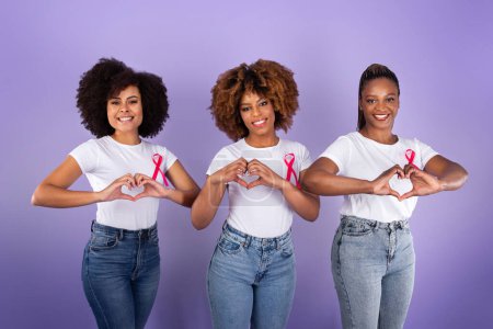 Foto de Oncología. Tres señoras jóvenes negras posando con cintas de cáncer de mama rosa y gestos en forma de corazón con los dedos de pie sobre fondo púrpura, sonriendo a la cámara. Concepto del Mes de Concientización - Imagen libre de derechos