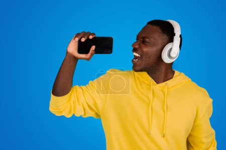 Foto de Feliz joven negro en auriculares inalámbricos canta en el teléfono inteligente con pantalla vacía en micrófono imaginario, aislado sobre fondo azul, estudio. Karaoke para superestrella, diversión, música en tiempo libre - Imagen libre de derechos