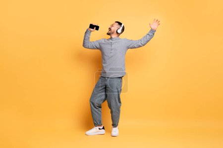 Téléchargez les photos : Portrait d'homme asiatique insouciant dans les écouteurs sans fil chantant avec Smartphone à la main, joyeux jeune homme utilisant le téléphone mobile comme microphone, s'amusant sur fond jaune, prise de vue pleine longueur - en image libre de droit