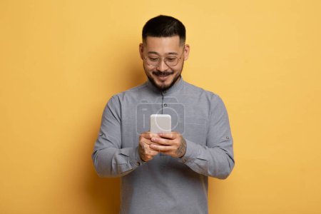 Foto de Sonriente joven asiático con teléfono inteligente en las manos posando sobre fondo de estudio amarillo, guapo chico milenario con tatuajes y gafas usando teléfono móvil para la comunicación en línea, espacio de copia - Imagen libre de derechos