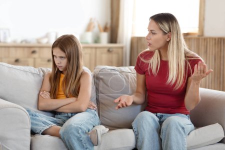 Photo for Adolescent misbehavior, disrespectful behaviour in pre-teens and teenagers. Blonde woman mother scolding her stubborn daughter at home, teen girl child sitting on couch with arms crossed next to mom - Royalty Free Image