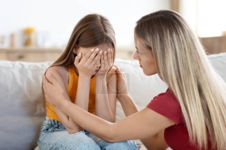 Loving supportive blonde woman mother supporting crying daughter, home interior, copy space. Mom and child girl teenager have quarrel, parent comforting upset kid. Parents and children conflicts