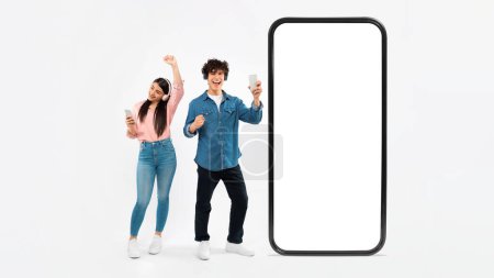Photo for Mobile Music Application. Cheerful Young Couple With Smartphones And Headphones Listening And Singing Songs, Dancing Near Huge Cellphone With Big Blank Screen, White Studio Background, Panorama - Royalty Free Image