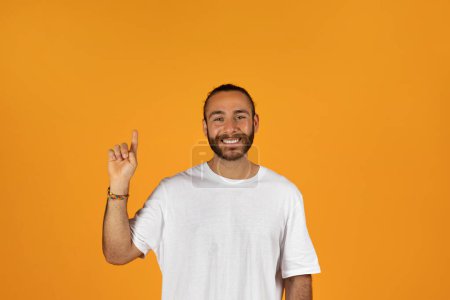 Photo for Positive millennial caucasian guy with beard in white t-shirt point finger up at empty space, isolated on orange studio background. Recommendation ad and offer, idea, advice to sale, work, study - Royalty Free Image