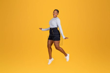 Photo for Excited attractive young African American woman in stylish outfit jumping in the air, smiling at camera isolated on yellow studio background. Happy black lady share positive emotions, copy space - Royalty Free Image
