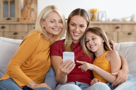 Photo for Cheerful loving family three generations of women mother, daughter and granny sitting on couch together, using phone at home, checking shopping application, scrolling, sharing interests - Royalty Free Image