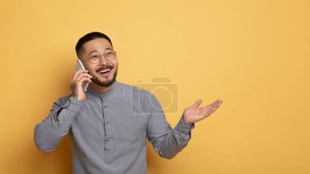 Téléchargez les photos : Communication mobile. excité jeune asiatique parler sur cellulaire et regarder loin, sourire Millennial Guy profiter agréable conversation tout en se tenant sur fond jaune, Copier l'espace - en image libre de droit