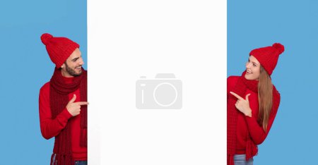 Photo for Place For Ad. Cheerful Young Couple In Winter Hats Pointing At White Advertisement Board, Happy Man And Woman Showing Free Copy Space For Your Xmas List, Standing On Blue Studio Background, Panorama - Royalty Free Image